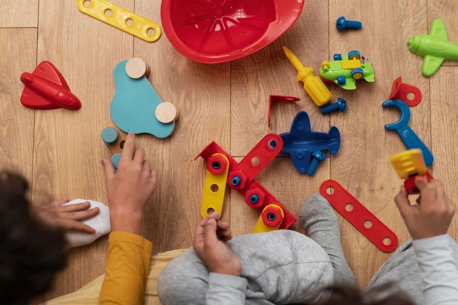Overhead view of two children sitting on a wooden floor surrounded by colourful toy construction pieces