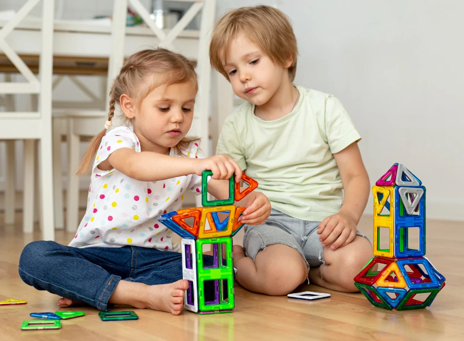 Two children sitting on the floor building a tower together from colourful magnetic tiles